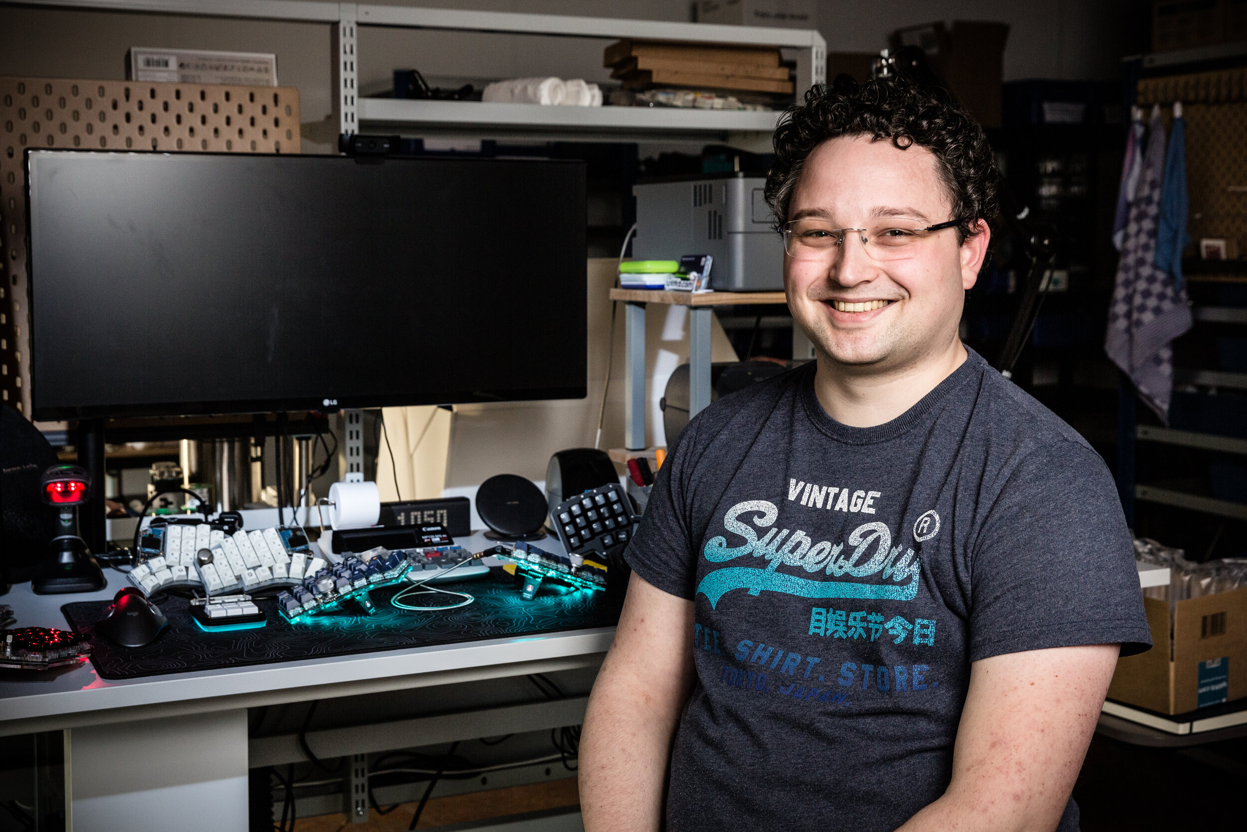 Thomas at his desk, where he displays several of the keyboards he has made. The keyboard with the glow is his daily driver: the Kyria, with a prototype of the tenting puck which allows him to position his keyboard halves at the angle he wants.